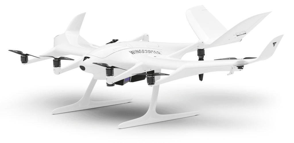 - RobotsInc.com White Wingcopter delivery drone on its landing gear against a white background, showing multiple propellers and a sleek fuselage.