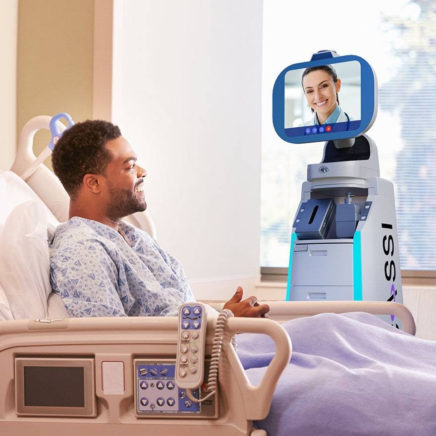 - RobotsInc.com Patient in a hospital bed smiles as a blue and white telepresence robot with a screen displaying a doctor approaches from the side.