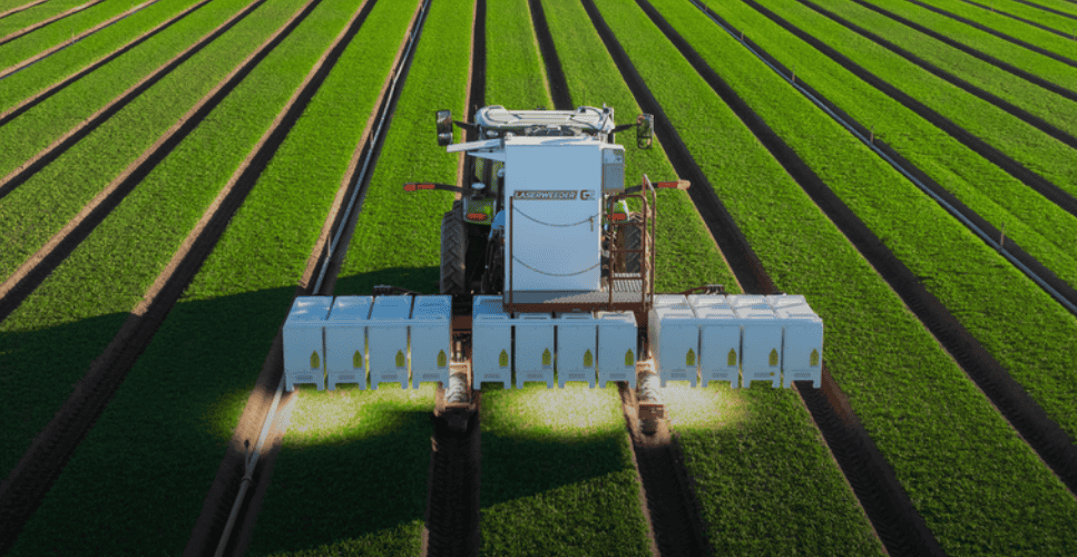 - RobotsInc.com Aerial view of a white agricultural machine planting crops in long, evenly spaced green rows with attached seedling trays in front of the machine.