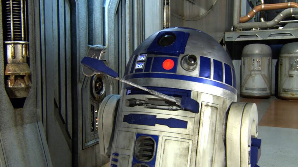 - RobotsInc.com R2-D2-like astromech droid in a high-tech maintenance bay with metal walls and pipes, leaning to the side with a blue tool arm extension nearby.