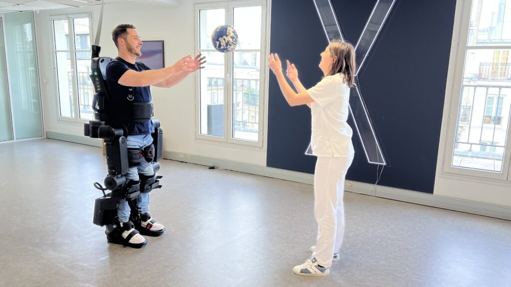 - RobotsInc.com Man in a powered exoskeleton suit reaching to catch a blue patterned ball from a woman in white in a bright studio space.