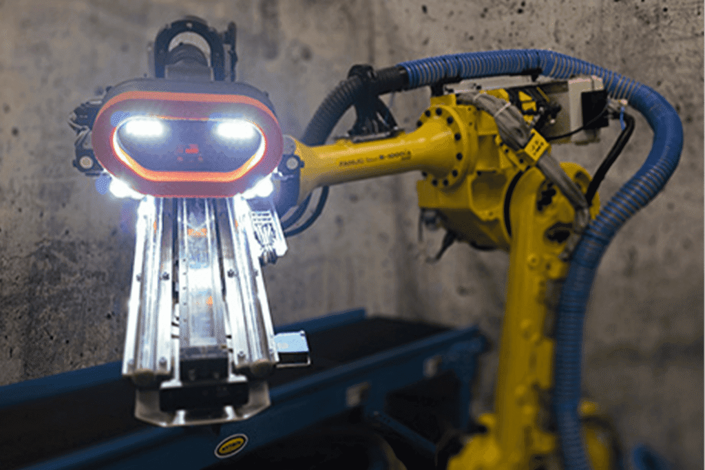 - RobotsInc.com Yellow industrial robotic arm with blue tubing, gripping a metal fixture on a workbench in a workshop setting.