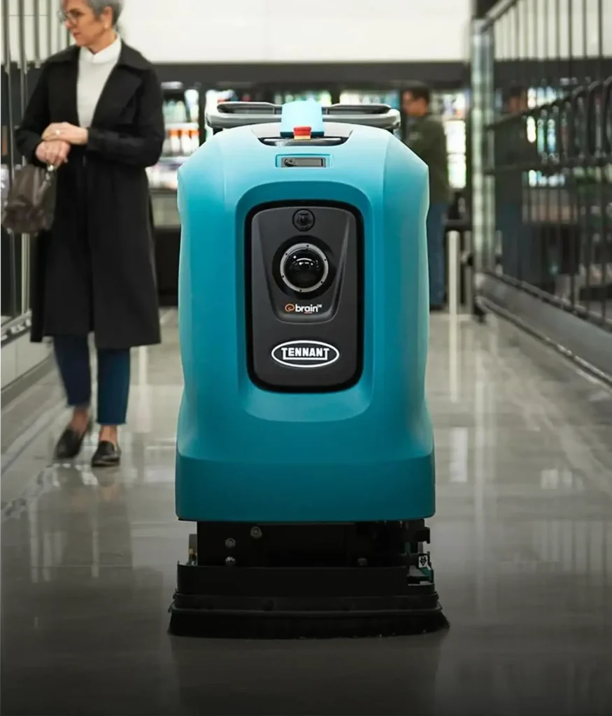 - RobotsInc.com Teal Tennant autonomous floor scrubber moved along a glass-walled corridor, with a person in a black coat standing nearby on the left.