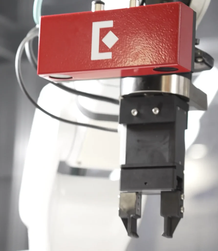 - RobotsInc.com Red rectangular industrial device with a white geometric logo mounted on a robotic arm, with black cables nearby.