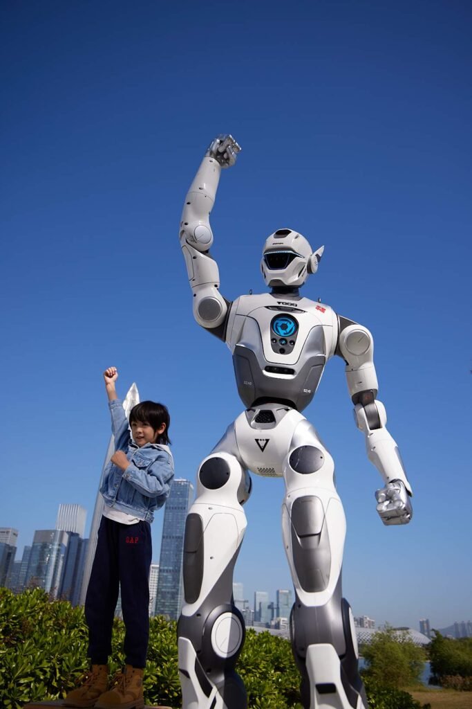 - RobotsInc.com A young boy standing beside a tall white humanoid robot outdoors, both raising a fist against a blue sky and city skyline behind them.