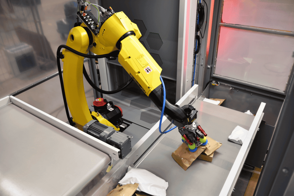 - RobotsInc.com Yellow industrial robotic arm inside a gray enclosure with visible cables and tooling, ready for testing or assembly work.