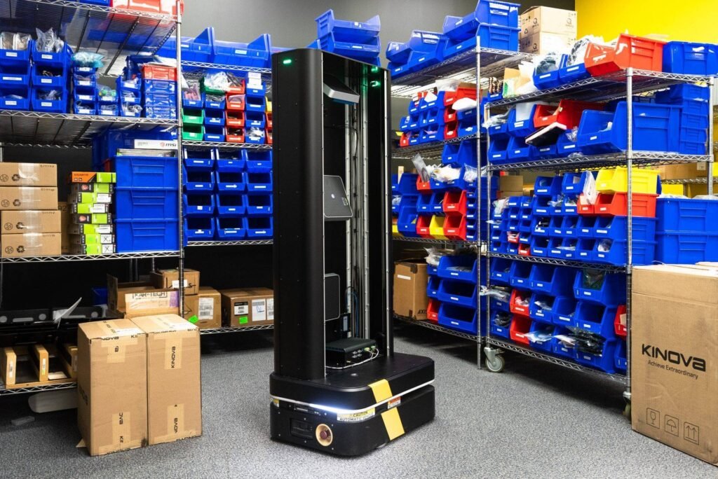 - RobotsInc.com Autonomous warehouse robot standing among shelves full of blue bins and boxes for inventory picking.