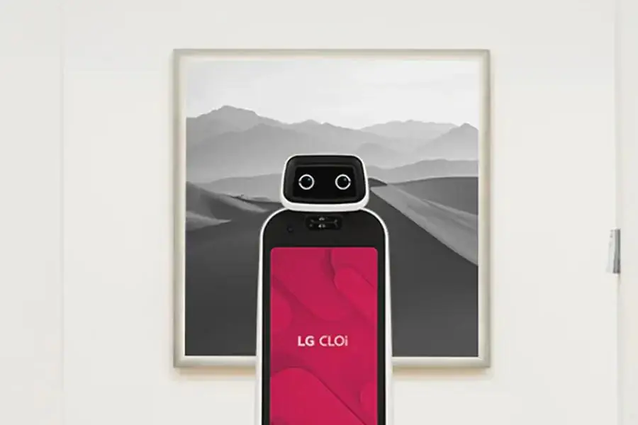 - RobotsInc.com LG CLOi service robot with a pink screen stands upright in front of a framed grayscale mountain landscape painting on a white wall.