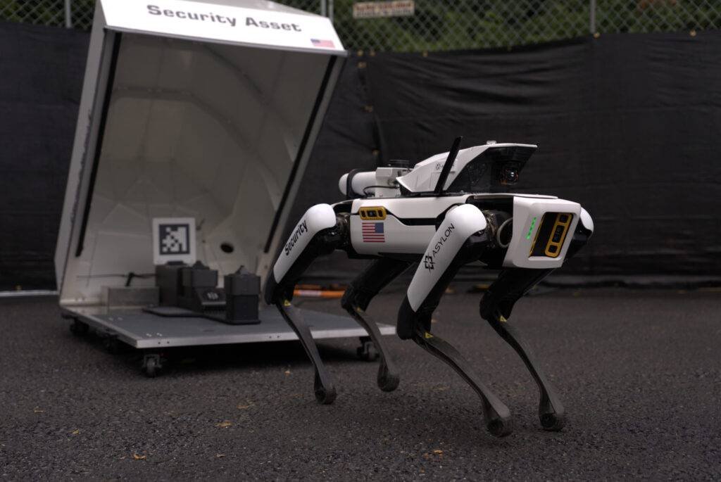 - RobotsInc.com Quadruped security robot walking on pavement beside a white portable security station labeled 'Security Asset'
