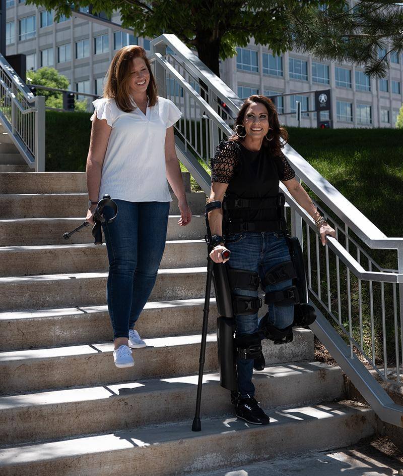 - RobotsInc.com Two women walk down outdoor stairs together; the woman on the right uses leg braces and crutches for support while the woman on the left smiles in a white blouse and jeans.