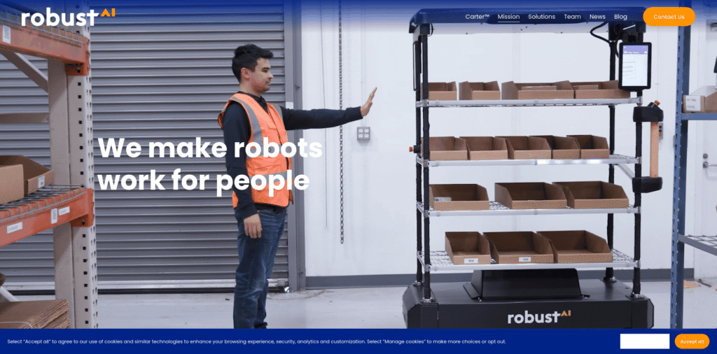 - RobotsInc.com Warehouse worker in an orange safety vest waves near a tall autonomous robot cart in a storage area with shelves behind him