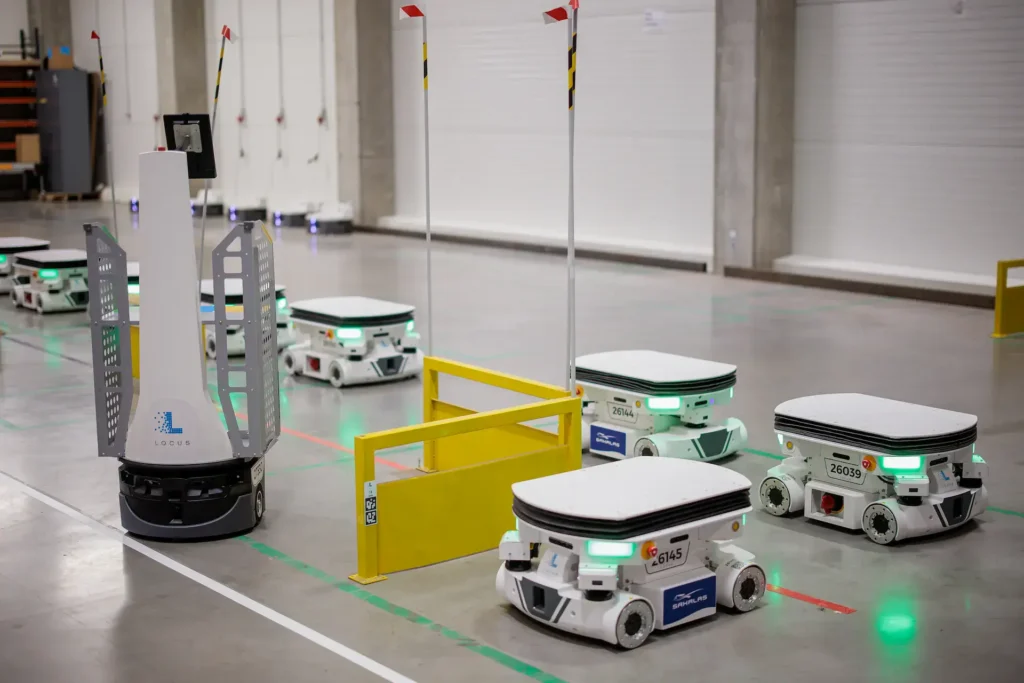 - RobotsInc.com Autonomous warehouse robots with green status lights navigating a clean industrial space near a yellow safety barrier.