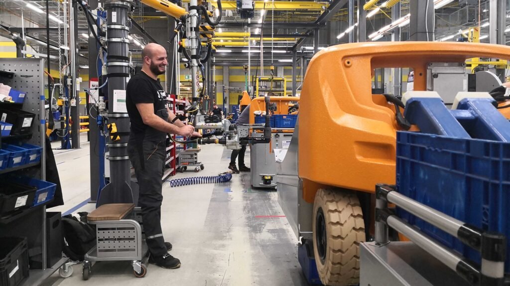 - RobotsInc.com Technician in black overalls smiling as he operates machinery on a busy factory floor, with an orange industrial vehicle and blue bins nearby.