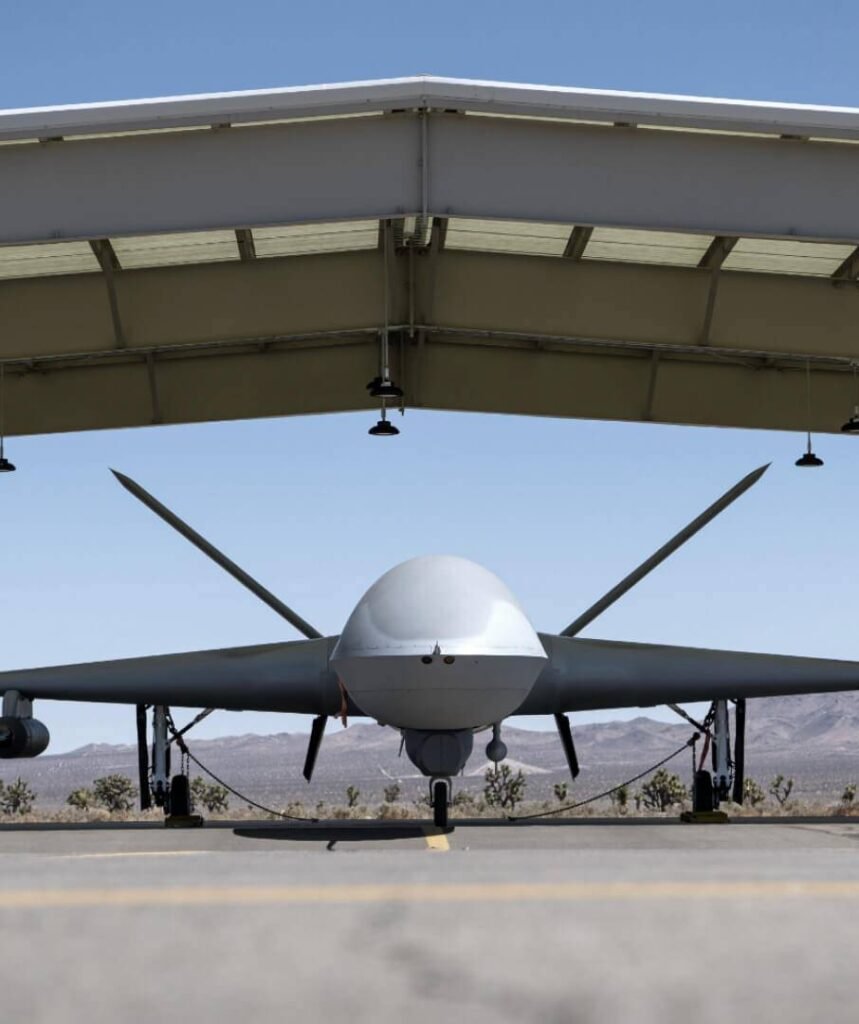 - RobotsInc.com Front view of a large unmanned aerial vehicle (drone) parked on a desert airstrip under a shelter, facing the camera with wings extended.