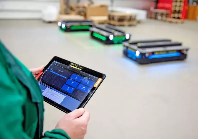 - RobotsInc.com Person holding a tablet controlling autonomous warehouse carts with blue/green lights in the background.