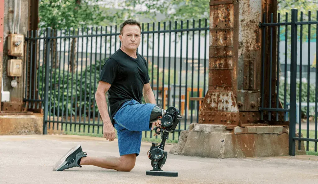 - RobotsInc.com Man kneels on one knee outdoors, wearing a black T-shirt and blue shorts, showing a mechanical leg prosthesis.