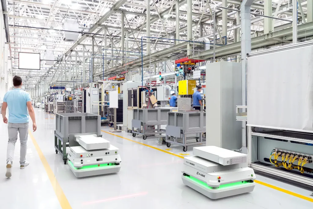 » RobotsInc.com Warehouse floor with wheeled autonomous robots moving along yellow lines as workers operate nearby machinery.