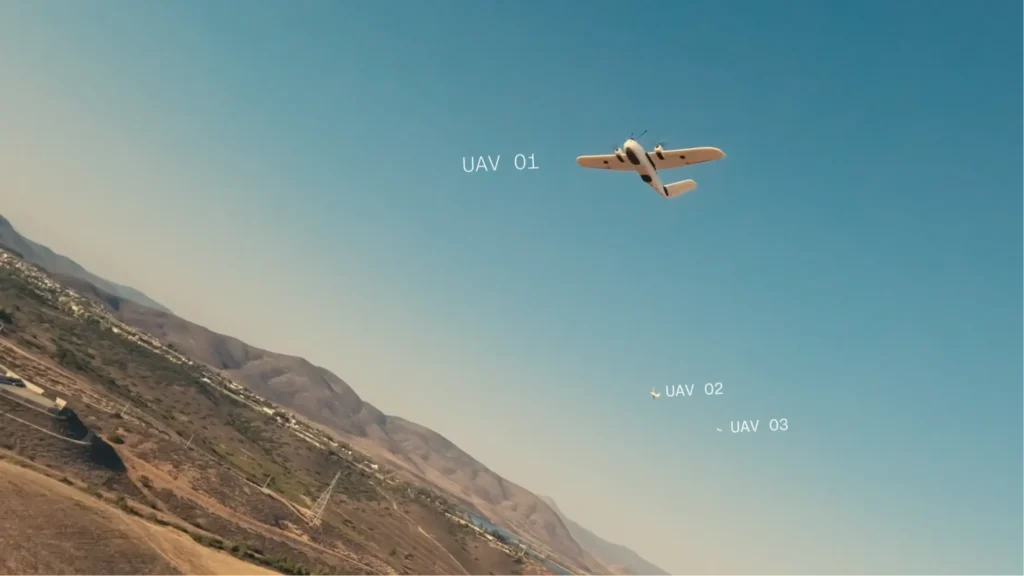 - RobotsInc.com A small white unmanned aerial vehicle flies over a desert valley with distant mountains and a blue sky; 'UAV 01' is visible in the scene.