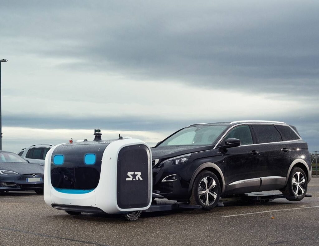 - RobotsInc.com Autonomous white and black robot shuttle attached to a black SUV in a parking lot, overcast sky above.