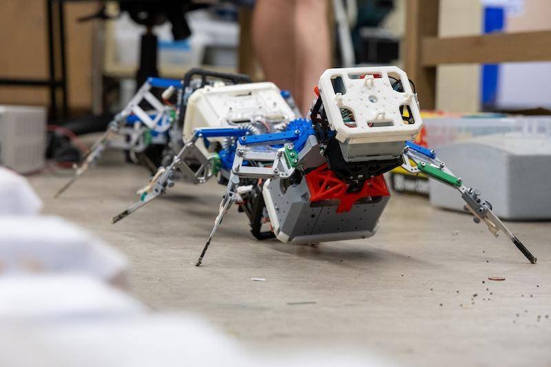 - RobotsInc.com Colorful modular legged robot crawling on a concrete workshop floor indoors.
