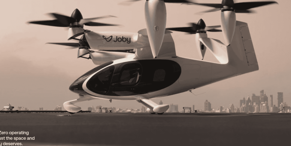 - RobotsInc.com Electric eVTOL aircraft by Joby moments before takeoff on a rooftop, city skyline in the background