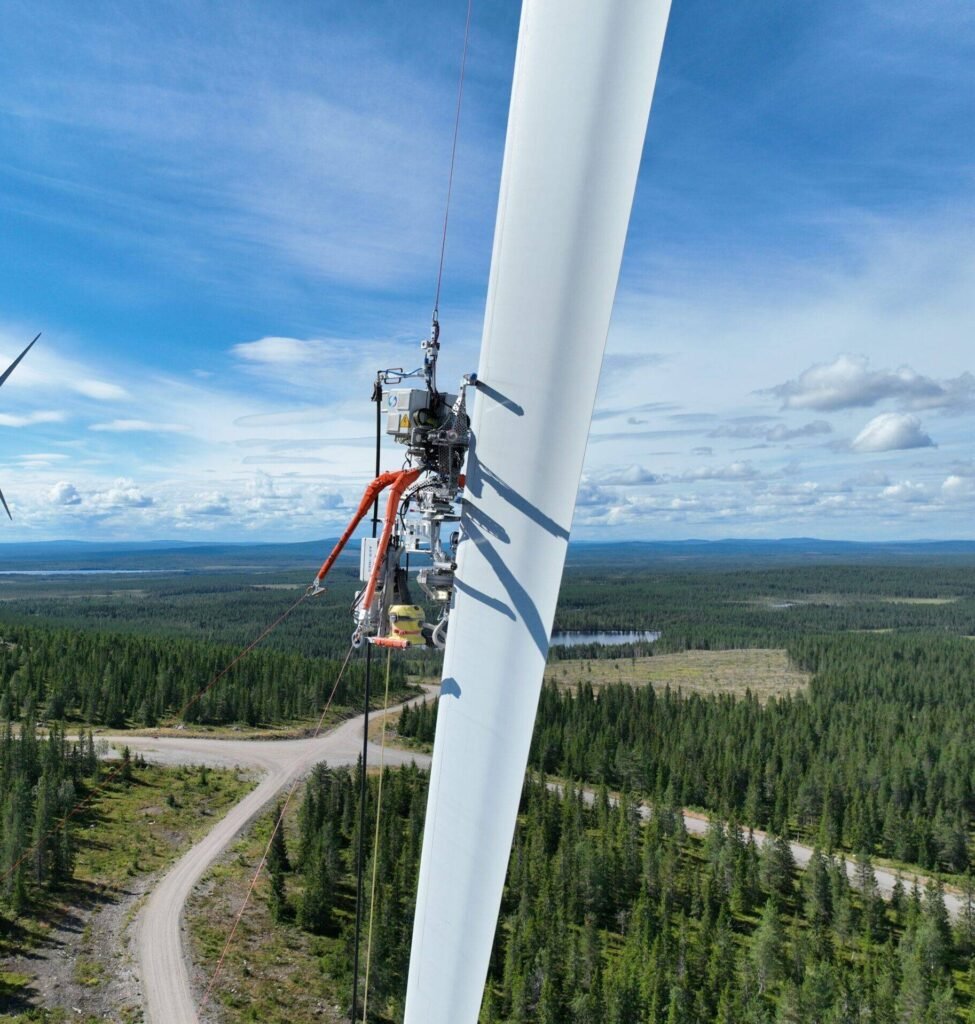 - RobotsInc.com Close-up view of a wind turbine blade with mounted maintenance equipment against a clear blue sky and forested landscape below.