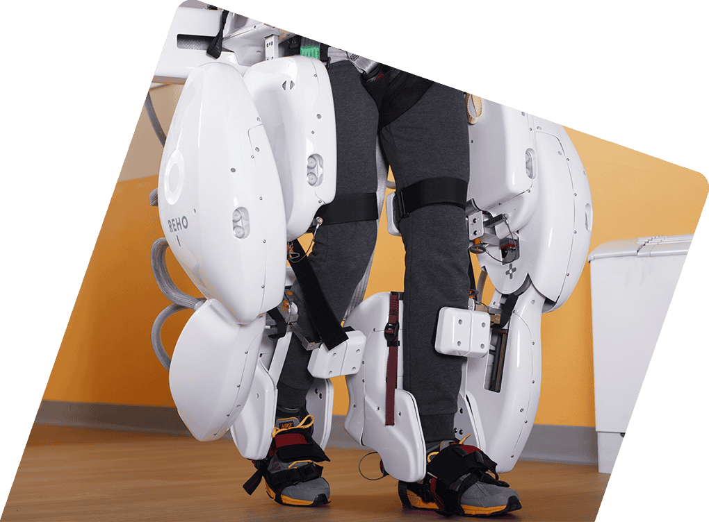 - RobotsInc.com Person wearing a white robotic exoskeleton on both legs, with straps and harnesses in a clinical setting.