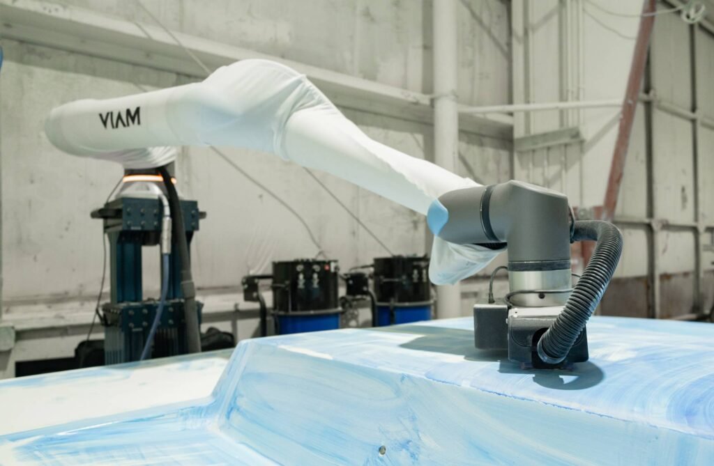 - RobotsInc.com Robotic arm applying light blue protective wrap to a car body in a factory setting, with hoses and machinery nearby.