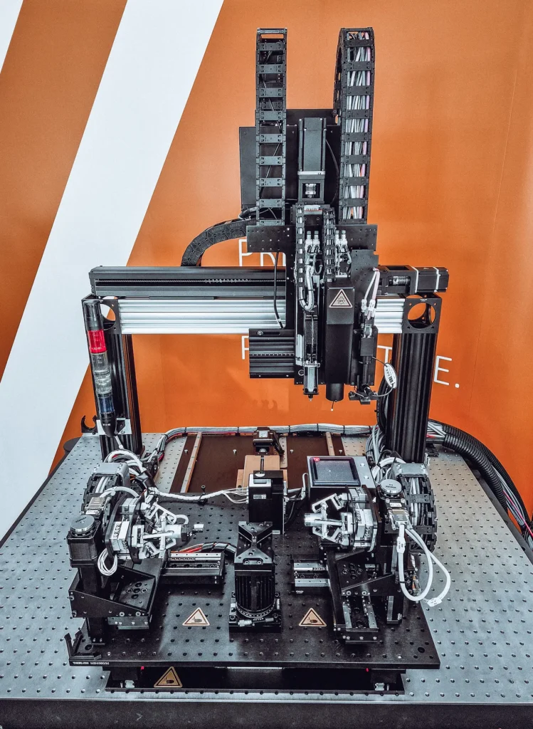 - RobotsInc.com Industrial robotic gantry with dual vertical rails and many cables on a perforated worktable against an orange wall