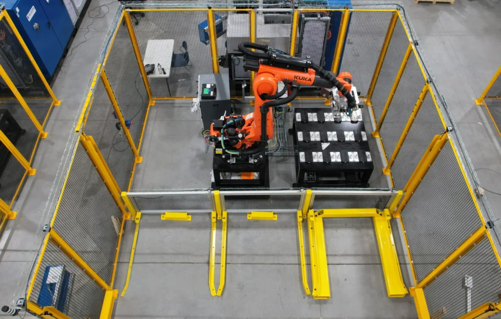 - RobotsInc.com Overhead view of an orange KUKA robotic arm inside a yellow safety cage, performing work on a bench.