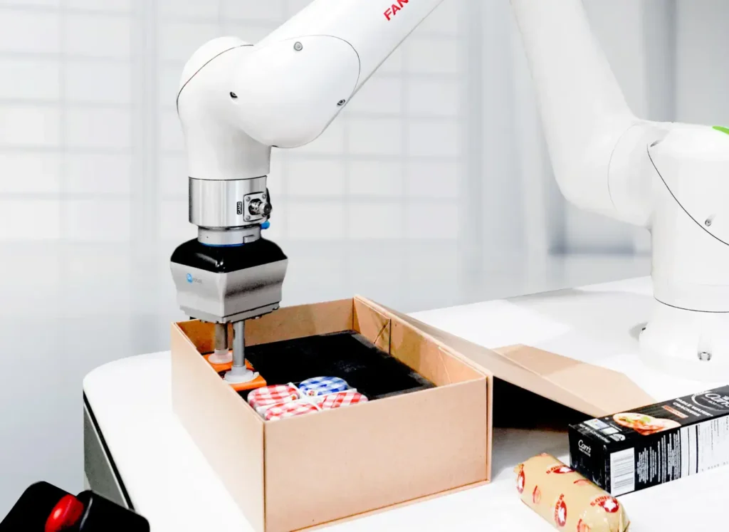 - RobotsInc.com White robotic arm placing snacks from a cardboard box on a lab table, showing automated packaging handling.