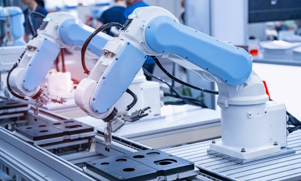 - RobotsInc.com Two blue robotic arms operate on a modern production line in a bright factory setting.