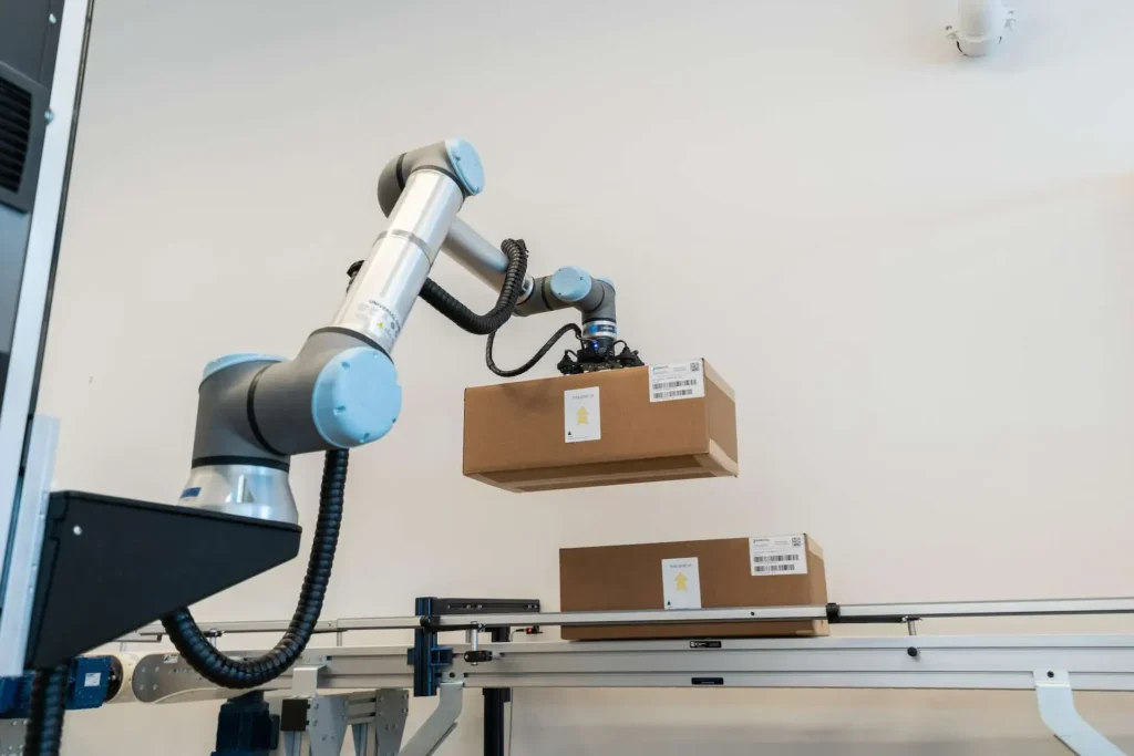 - RobotsInc.com Collaborative robotic arm lifting a brown box on a conveyor system in a warehouse setting.