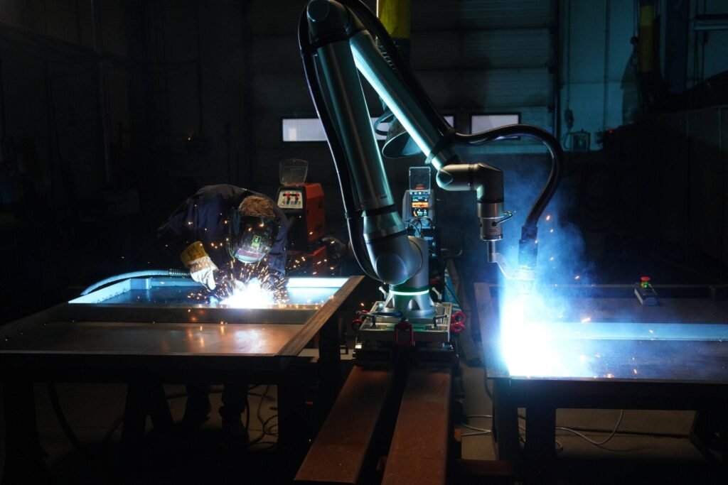 - RobotsInc.com Robotic welding arm at work, sparks flying over a metal workbench in a dim workshop.