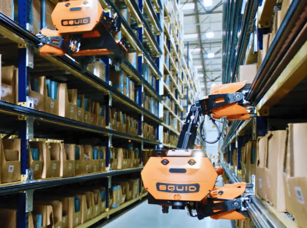 - RobotsInc.com Autonomous orange warehouse robot moving between tall shelves filled with boxes and pallets.