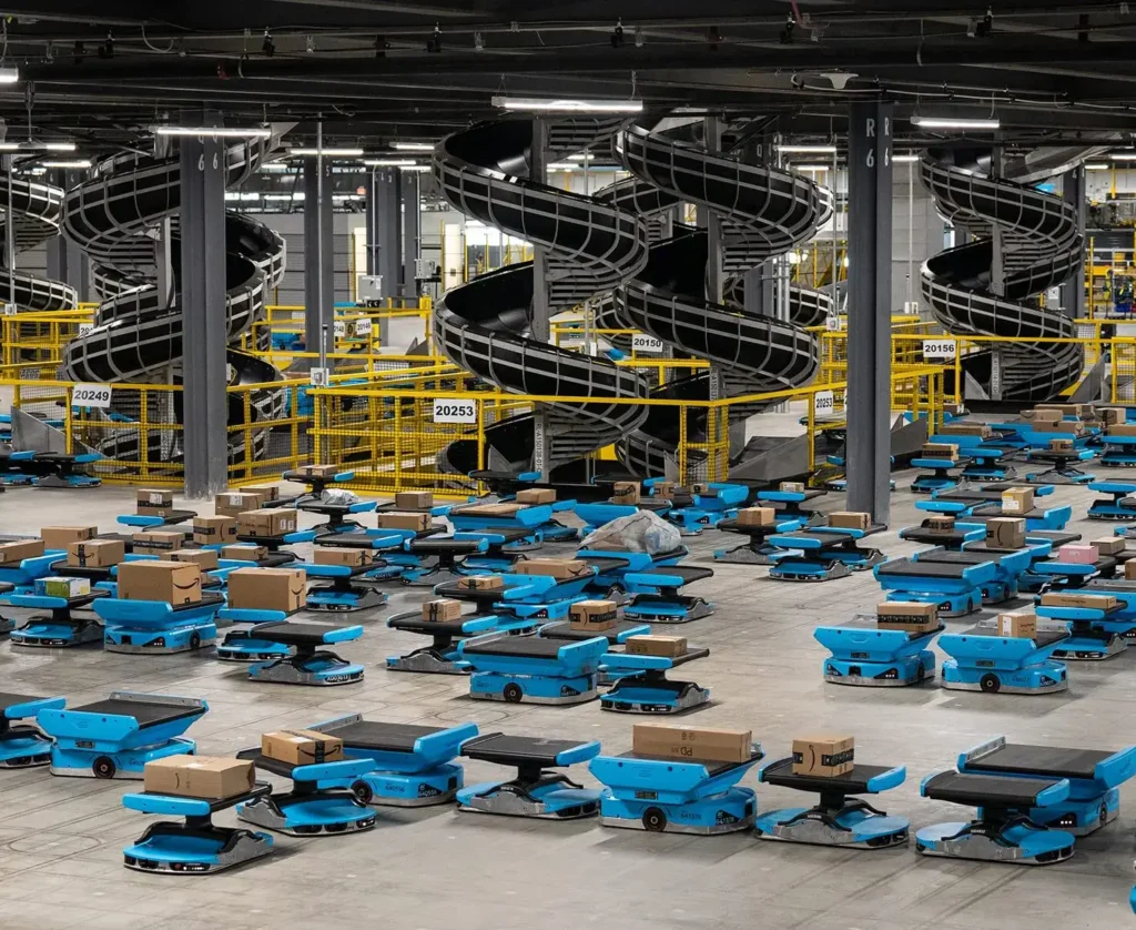 - RobotsInc.com Warehouse filled with blue autonomous delivery robots and scattered cardboard boxes, with twisting black conveyor slides in the background.