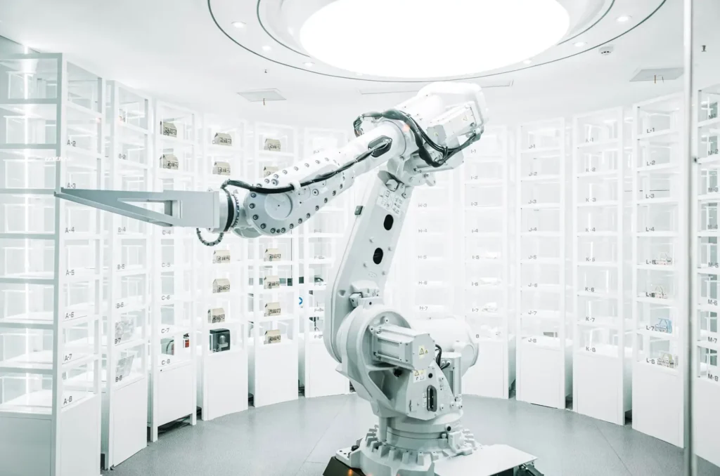- RobotsInc.com Industrial robotic arm operating in a high-tech storage room with labeled rows of bins and shelves lining the space