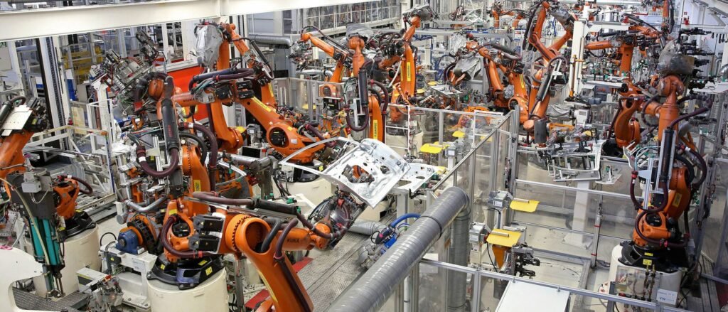 - RobotsInc.com Industrial robot assembly line with numerous orange robotic arms working on metallic car frames in a factory setting.