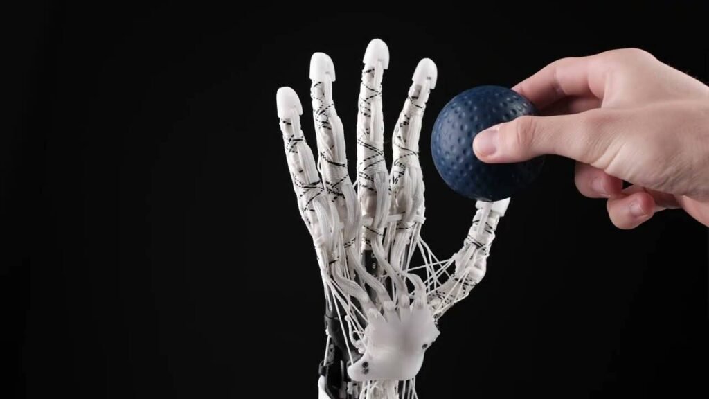 - RobotsInc.com Robotic prosthetic hand with a human hand pressing a blue textured ball against a black background.