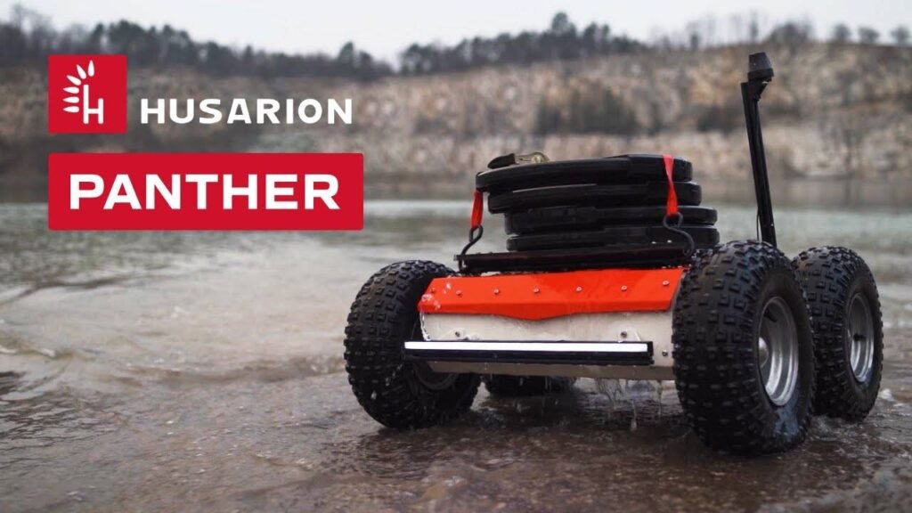- RobotsInc.com Promotional image for Husarion Panther robotic rover on a muddy shore with large knobby tires and a stacked module on top.