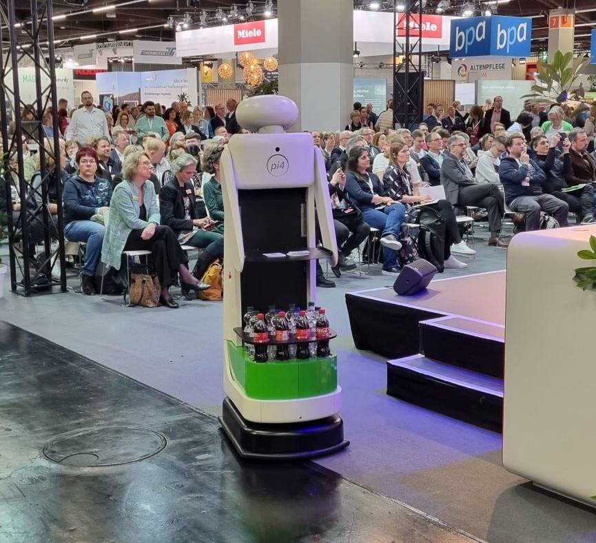 - RobotsInc.com A service robot carrying bottles on a green tray at a conference stage while an audience watches the presentation.