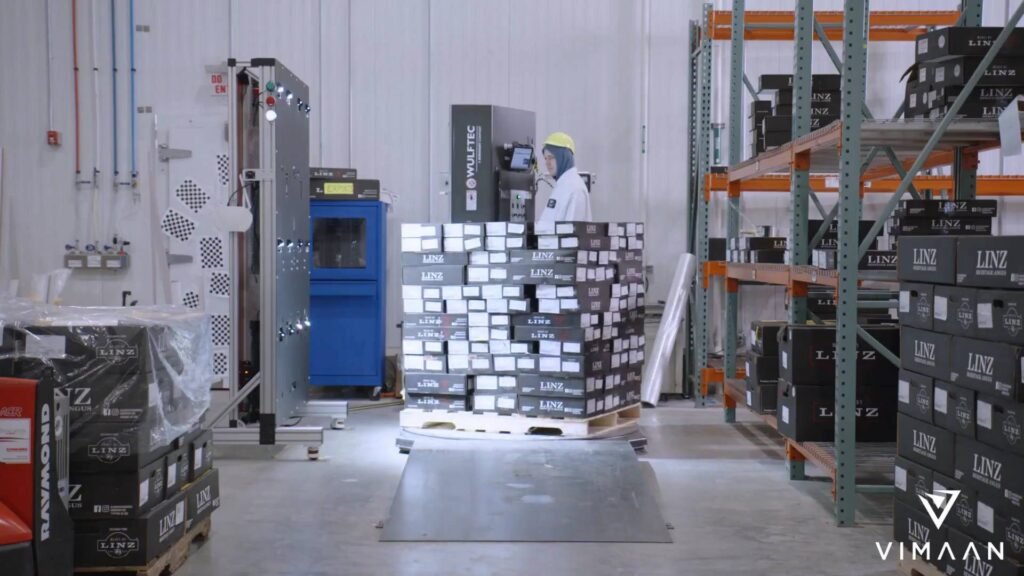 - RobotsInc.com Warehouse worker in yellow hard hat operates control panel beside stacked LINZ boxes on pallets and metal shelves nearby.