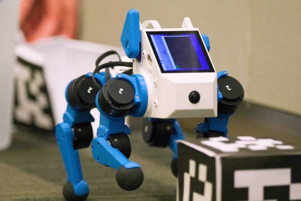 - RobotsInc.com Small blue-and-white four‑legged robot with a touchscreen head on a lab bench, facing a black-and-white cube.