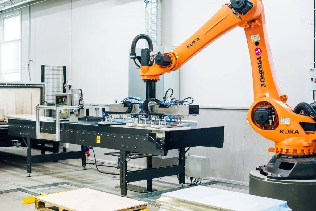 - RobotsInc.com Industrial robotic arm (KUKA) operates over a metal conveyor table in a factory setting.
