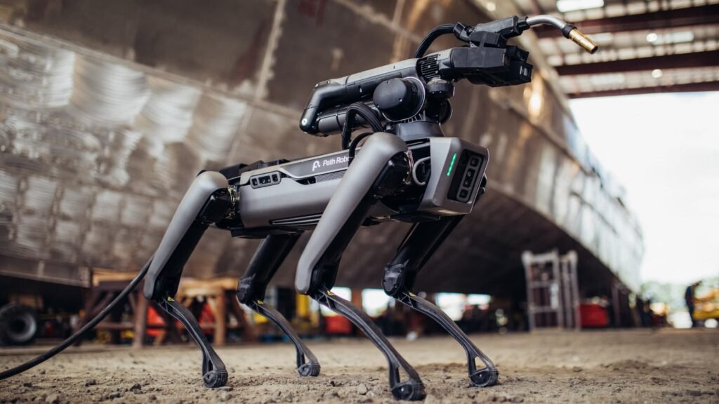 - RobotsInc.com Quadruped industrial robot on a dusty factory floor with cables and machinery in the background.