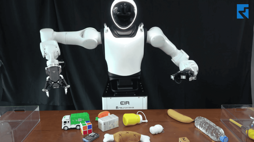 - RobotsInc.com White humanoid robot with articulated arms stands behind a table, with colorful blocks and miscellaneous objects in the foreground against a dark backdrop.