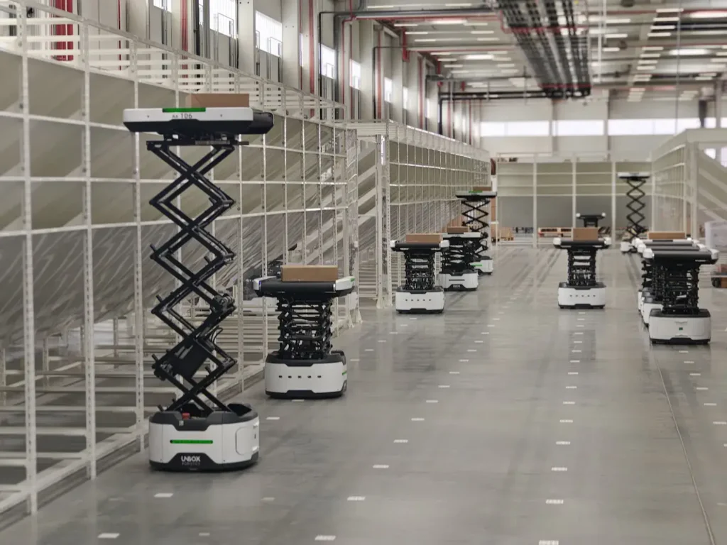 » RobotsInc.com Autonomous delivery robots with scissor-lift platforms carrying packages in a spacious warehouse aisle under bright overhead lights.