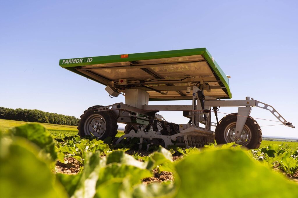 » RobotsInc.com Green agricultural machine on tracks in a field, with leafy crops in the foreground and a clear blue sky above.