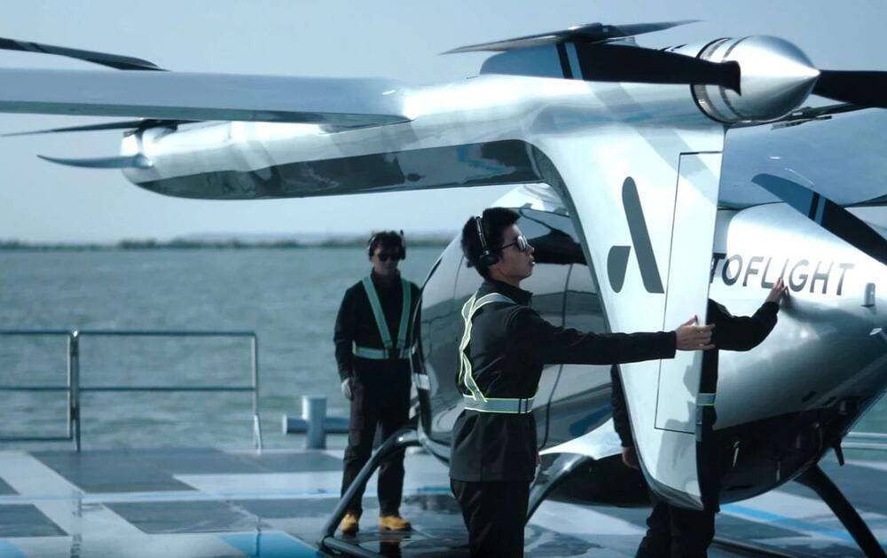 - RobotsInc.com Two crew members in safety vests guide a white futuristic aircraft on a marina deck by the water, one with arms extended to station the craft.