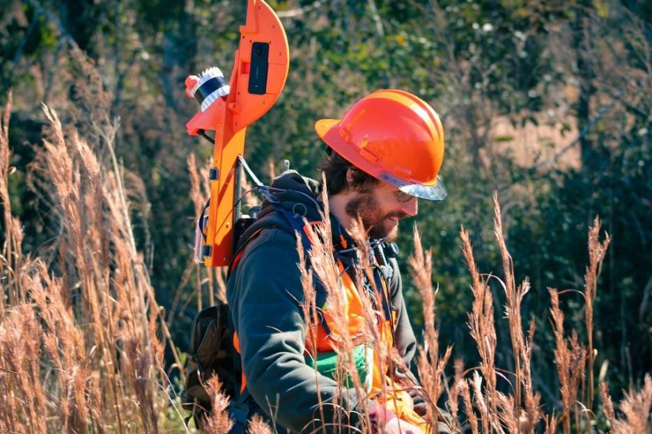 » RobotsInc.com Male worker in an orange hard hat and safety vest walks through tall dried grass with surveying equipment strapped to his back.